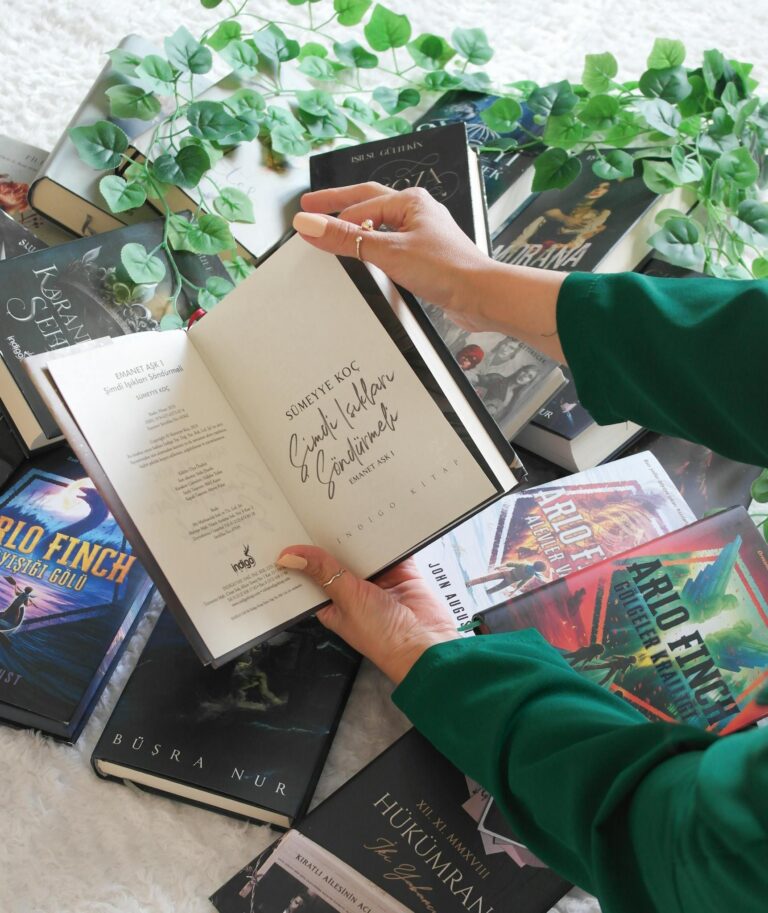 Person holding open book with signature among stacked novels and greenery.