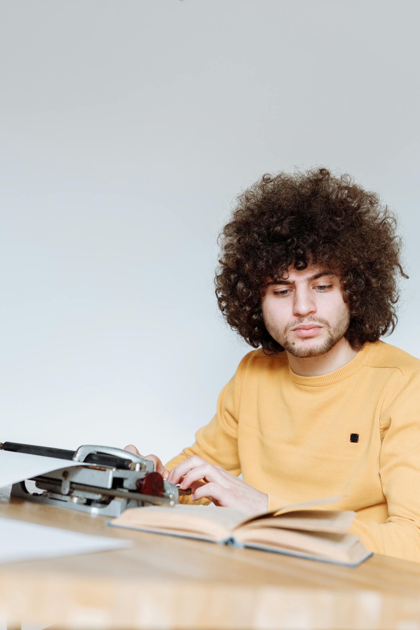 Young man with curly hair types on vintage typewriter while reading an open book at a wooden desk.
